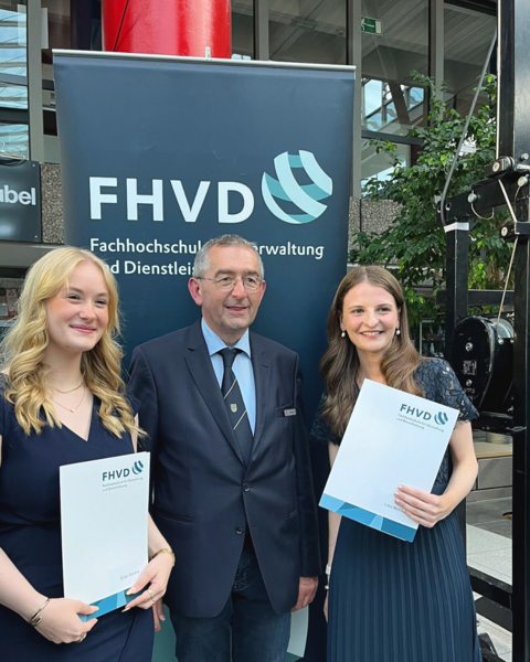 Norbert Krüger (Ausbildungsverantwortlicher) mit den Absolventinnen Eilien Sievers (l.) und Jule Böttcher vor einem Banner der FHVD