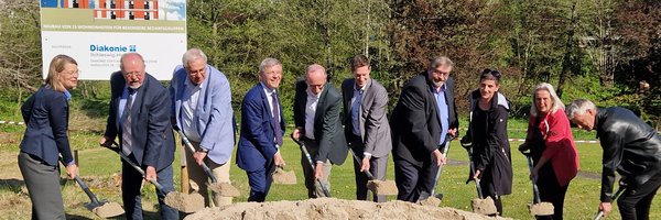 Spatenstich für ein zukunftsweisendes Bauprojekt im Ansgarweg 4–5: Vertreter*innen der Diakonie Stiftung Schleswig-Holstein, des Landes S-H, des Kreises Schleswig-Flensburg und der Stadt Schleswig. Alle Personen haben einen Spaten in der Hand und stechen in den Sandhaufen vor ihnen. Im Hintergrund Bauschild und Bäume