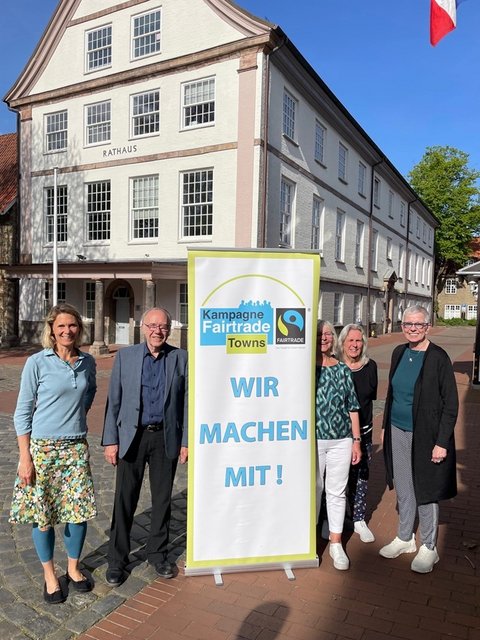 Das Foto zeigt aus dem Steuerungsgruppen-Team Sina Clorius (Ratsversammlung), Matthias Hertel und Ute Ulrich (Verein zur Förderung des fairen Handels Schleswig e. V.) Maren Korban und Inke Asmussen (beide Ratsversammlung) mit einem Aufsteller "Wir machen mit" vor dem Schleswiger Rathaus